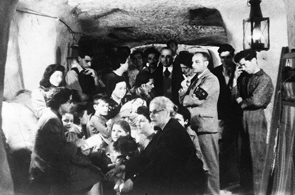 1942年5月，瓦萊塔空襲期間的避難所內，成人與孩子聚在岩壁之間，壁上的燈照着擁擠的小室。照片由英國皇家空軍航空史部提供。